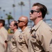 A group of military personnel in tan/khaki uniforms are standing in formation, likely during a ceremony or official event. The individuals are wearing U.S. Navy Service Uniform (Type III or Summer Whites variant) adorned with ribbons, badges, and insignia. The setting appears to be an outdoor location with palm trees visible in the background, suggesting a warm-climate naval installation.