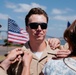 A young man in a tan/khaki military uniform is having insignia or rank devices pinned onto his collar by two individuals — one in a military uniform with a peaked cap, and one in civilian clothing. An American flag is prominently displayed in the background, along with what appears to be a waterfront or harbor setting. This appears to be a military promotion or commissioning ceremony.