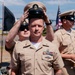 A U.S. Navy promotion or pinning ceremony is taking place outdoors, likely at a naval base near a waterfront. A chief petty officer's cover (hat) is being placed on the head of a sailor in the foreground, a traditional part of the Chief Petty Officer initiation or promotion ceremony. An American flag and what appears to be the Coronado Bridge in San Diego are visible in the background.