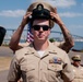 A U.S. Navy Chief Petty Officer is having his combination cover (cap) placed on his head during what appears to be a Chief Petty Officer pinning or advancement ceremony. He is wearing the tan Service Uniform (SU) with multiple ribbons, warfare devices, and collar insignia. The outdoor ceremony appears to take place near a waterfront, likely a naval installation in San Diego based on the visible bridge in the background.