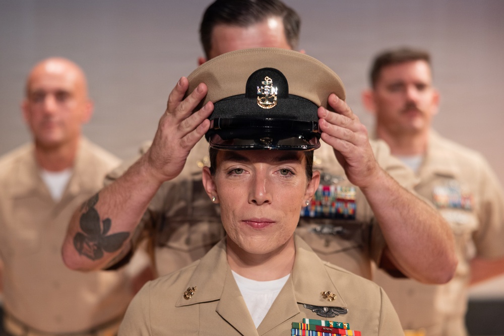A U.S. Navy Chief Petty Officer promotion ceremony is taking place, with a senior enlisted member placing the distinctive khaki combination cover (cap) on the newly promoted sailor's head. The central figure is a female sailor in Service Khaki uniform adorned with multiple ribbons, warfare devices, and collar insignia. Two other sailors in the same uniform are visible in the background, slightly out of focus.