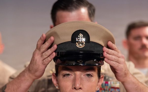 A U.S. Navy Chief Petty Officer promotion ceremony is taking place, with a senior enlisted member placing the distinctive khaki combination cover (cap) on the newly promoted sailor's head. The central figure is a female sailor in Service Khaki uniform adorned with multiple ribbons, warfare devices, and collar insignia. Two other sailors in the same uniform are visible in the background, slightly out of focus.