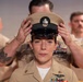 A U.S. Navy Chief Petty Officer promotion ceremony is taking place, with a senior enlisted member placing the distinctive khaki combination cover (cap) on the newly promoted sailor's head. The central figure is a female sailor in Service Khaki uniform adorned with multiple ribbons, warfare devices, and collar insignia. Two other sailors in the same uniform are visible in the background, slightly out of focus.