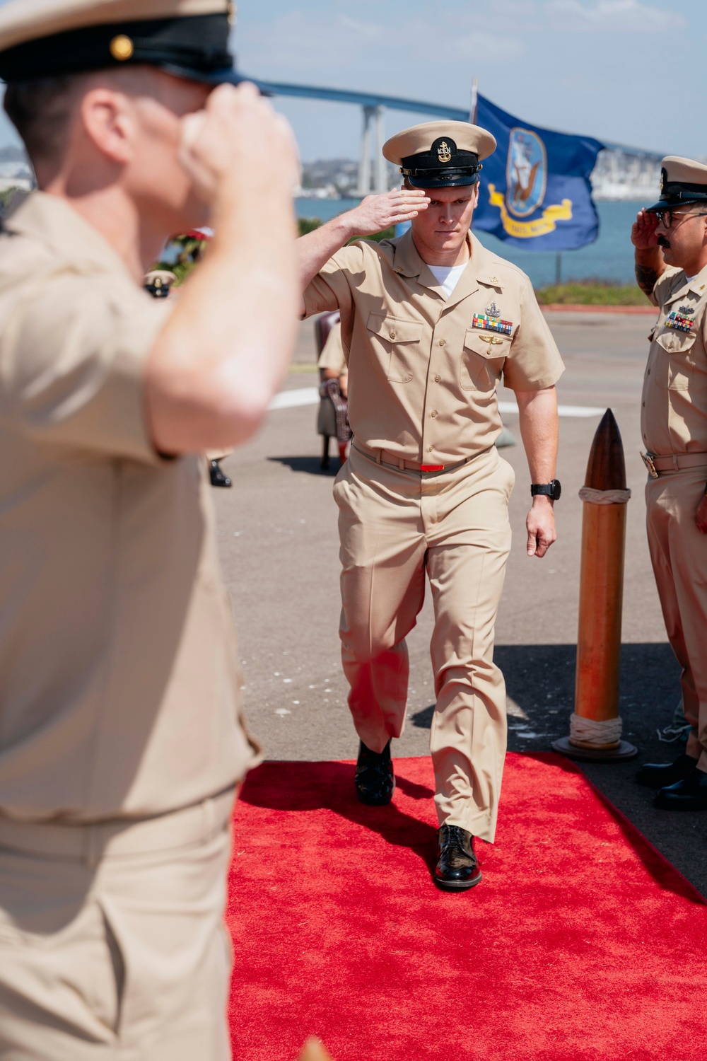 A U.S. Navy military ceremony is taking place outdoors, likely at a naval installation near San Diego, California, identifiable by the Coronado Bridge visible in the background. A senior enlisted sailor in Service Khaki uniform walks along a red carpet while exchanging salutes with fellow personnel. The scene appears to be a formal sideboy ceremony, consistent with a change of command or retirement event.