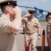 A U.S. Navy military ceremony is taking place outdoors, likely at a naval installation near San Diego, California, identifiable by the Coronado Bridge visible in the background. A senior enlisted sailor in Service Khaki uniform walks along a red carpet while exchanging salutes with fellow personnel. The scene appears to be a formal sideboy ceremony, consistent with a change of command or retirement event.