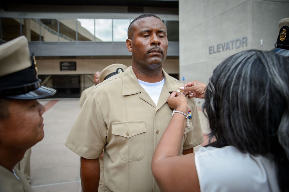A military ceremony is taking place outdoors near a building with an elevator sign. A woman is pinning a medal or insignia onto the uniform of a male Navy service member in a tan/khaki uniform. Other uniformed personnel are present in the background and to the left.