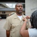 A military ceremony is taking place outdoors near a building with an elevator sign. A woman is pinning a medal or insignia onto the uniform of a male Navy service member in a tan/khaki uniform. Other uniformed personnel are present in the background and to the left.