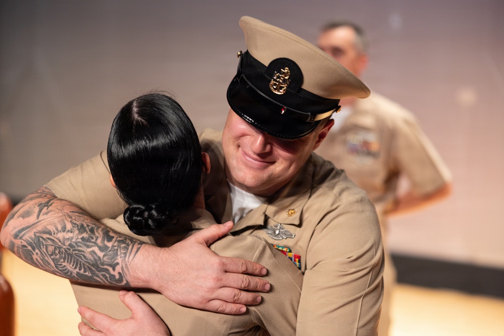 Two U.S. Navy personnel in tan service uniforms are sharing an emotional embrace, likely at a military ceremony such as a promotion or pinning event. The male sailor, wearing a chief petty officer combination cover with gold insignia, is smiling warmly while hugging a fellow service member with an extensively tattooed arm. A third uniformed individual is visible but out of focus in the background.