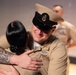 Two U.S. Navy personnel in tan service uniforms are sharing an emotional embrace, likely at a military ceremony such as a promotion or pinning event. The male sailor, wearing a chief petty officer combination cover with gold insignia, is smiling warmly while hugging a fellow service member with an extensively tattooed arm. A third uniformed individual is visible but out of focus in the background.