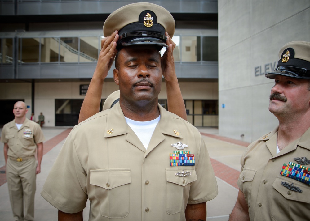 A U.S. Navy Chief Petty Officer is having his combination cover (cap) placed on his head during what appears to be a chief petty officer pinning or frocking ceremony. The central figure stands at attention with eyes closed while hands place the distinctive CPO cover on his head. Two other Navy personnel in khaki uniforms are visible in the scene, one closely observing and one in the background.