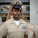 A U.S. Navy Chief Petty Officer is having his combination cover (cap) placed on his head during what appears to be a chief petty officer pinning or frocking ceremony. The central figure stands at attention with eyes closed while hands place the distinctive CPO cover on his head. Two other Navy personnel in khaki uniforms are visible in the scene, one closely observing and one in the background.