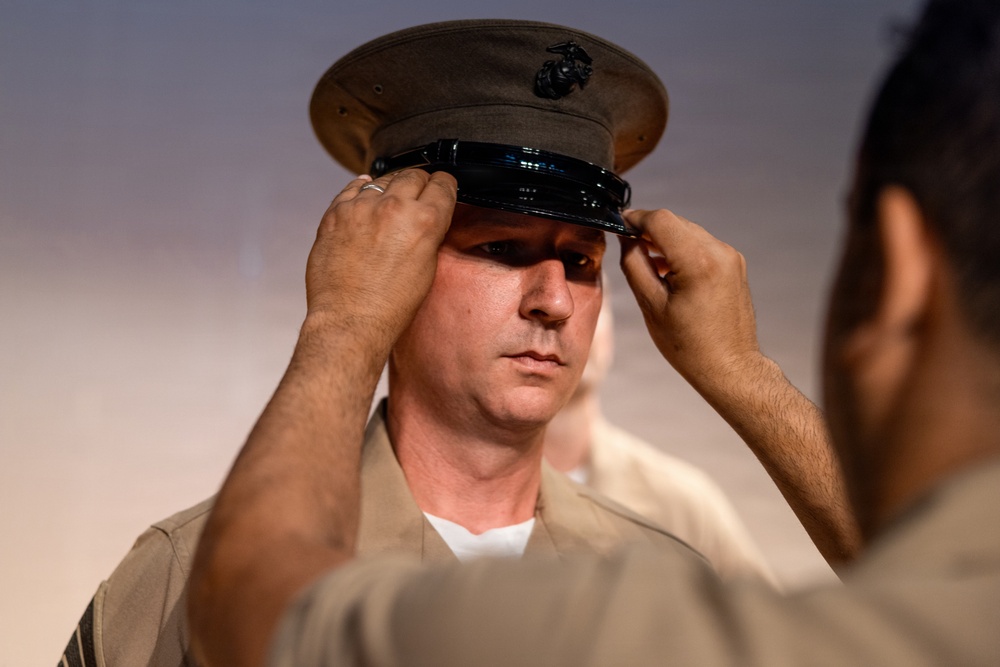 A U.S. Marine is having his dress cover (uniform cap) placed or adjusted on his head by another person. The cap features the distinctive Eagle, Globe, and Anchor emblem of the United States Marine Corps. Both individuals are wearing tan/khaki Marine Corps service uniforms.