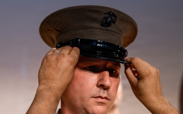 A U.S. Marine is having his dress cover (uniform cap) placed or adjusted on his head by another person. The cap features the distinctive Eagle, Globe, and Anchor emblem of the United States Marine Corps. Both individuals are wearing tan/khaki Marine Corps service uniforms.