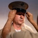 A U.S. Marine is having his dress cover (uniform cap) placed or adjusted on his head by another person. The cap features the distinctive Eagle, Globe, and Anchor emblem of the United States Marine Corps. Both individuals are wearing tan/khaki Marine Corps service uniforms.