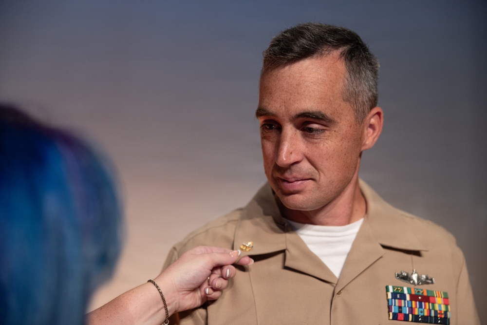 A U.S. Navy officer in tan service uniform (Service Khaki) is having a gold insignia or device pinned onto his uniform by another person whose hand is visible in the foreground. The officer displays multiple service ribbons and a warfare qualification badge on his chest. This appears to be a military promotion or award ceremony.