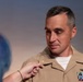 A U.S. Navy officer in tan service uniform (Service Khaki) is having a gold insignia or device pinned onto his uniform by another person whose hand is visible in the foreground. The officer displays multiple service ribbons and a warfare qualification badge on his chest. This appears to be a military promotion or award ceremony.