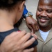 A U.S. Navy enlisted service member in tan/khaki uniform is smiling broadly while a person pins something onto his collar or shoulder area. The scene appears to be a military promotion or advancement ceremony. The service member displays multiple ribbons, warfare qualification badges, and chief petty officer insignia on his uniform.