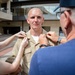 A military service member in a tan/khaki uniform is having insignia or rank devices pinned onto his collar by two individuals — a woman on his left and an older man on his right. This appears to be a military promotion or pinning ceremony taking place outdoors in front of a multi-story building. The service member stands at attention while family members or colleagues perform the traditional pinning.