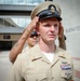 Two U.S. Navy personnel in Service Khaki uniforms are rendering a salute in front of what appears to be a military or government building. The foreground sailor wears a Chief Petty Officer combination cover with USN anchor insignia and displays multiple service ribbons, warfare devices, and collar devices on his uniform. A second sailor is visible in the background, also saluting.