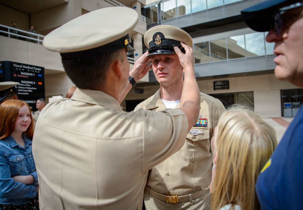 A military promotion or pinning ceremony is taking place outdoors at Naval Medical Center San Diego. One uniformed Navy service member is placing a chief petty officer cover (hat) on another service member's head, a traditional part of a CPO initiation or promotion ceremony. Family members, including children, are present as witnesses to the event.