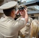A military promotion or pinning ceremony is taking place outdoors at Naval Medical Center San Diego. One uniformed Navy service member is placing a chief petty officer cover (hat) on another service member's head, a traditional part of a CPO initiation or promotion ceremony. Family members, including children, are present as witnesses to the event.