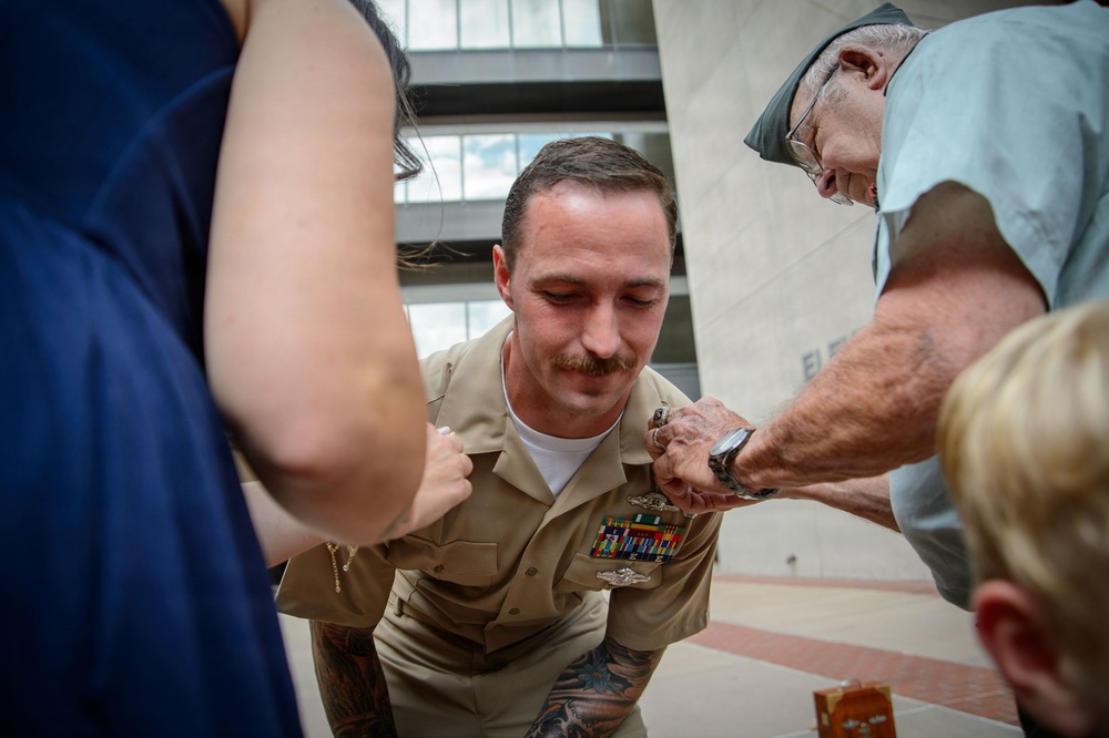 A U.S. Navy sailor in khaki service uniform is being pinned with a new insignia or rank device by an older civilian man, while others assist. The sailor is leaning forward slightly as multiple people participate in the pinning ceremony. This appears to be a military promotion or qualification ceremony taking place outdoors near a building.