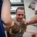 A U.S. Navy sailor in khaki service uniform is being pinned with a new insignia or rank device by an older civilian man, while others assist. The sailor is leaning forward slightly as multiple people participate in the pinning ceremony. This appears to be a military promotion or qualification ceremony taking place outdoors near a building.