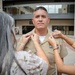 A U.S. Navy servicemember in khaki dress uniform is having collar devices or insignia pinned onto his uniform by two women, likely family members. This appears to be a promotion ceremony, a common military tradition where loved ones pin new rank insignia. The scene takes place outdoors in front of a modern building.