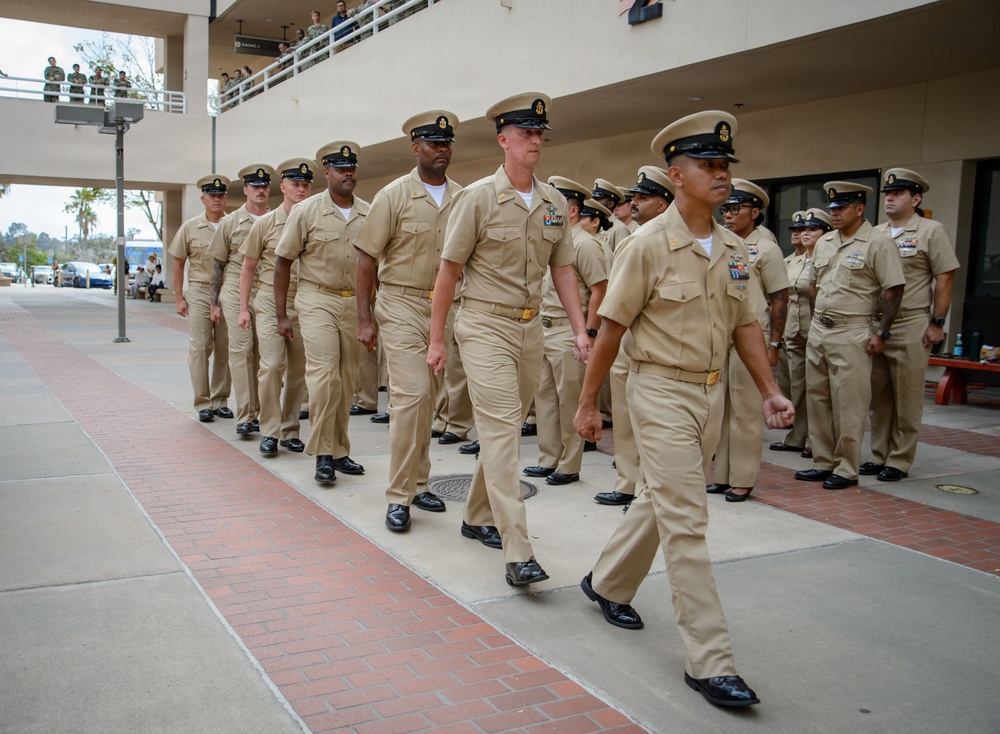 A formation of U.S. Navy enlisted personnel, predominantly Chief Petty Officers, are marching in formation at what appears to be a military installation. The personnel are wearing the standard khaki Service Uniform (Type II) with combination covers (garrison caps). Spectators can be seen observing from an elevated walkway in the background.