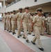 A formation of U.S. Navy enlisted personnel, predominantly Chief Petty Officers, are marching in formation at what appears to be a military installation. The personnel are wearing the standard khaki Service Uniform (Type II) with combination covers (garrison caps). Spectators can be seen observing from an elevated walkway in the background.