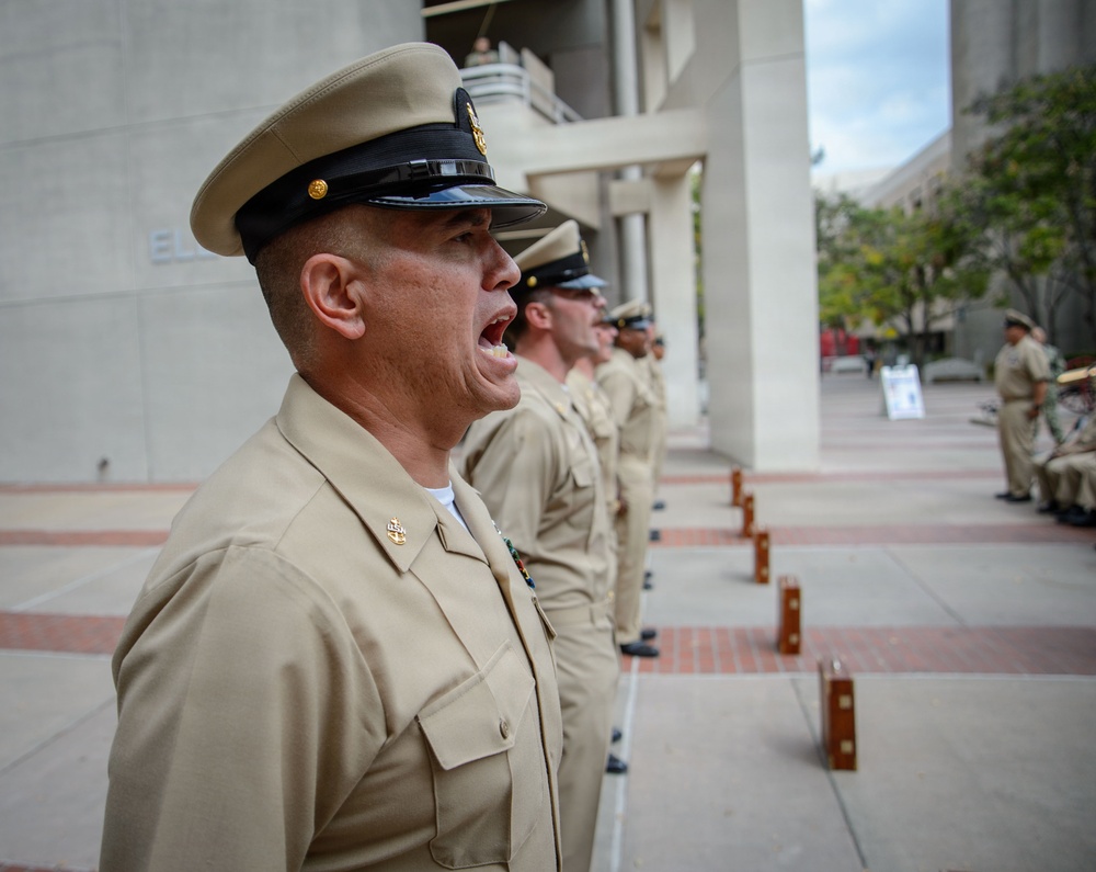 A group of U.S. Navy personnel in Service Khaki uniforms are standing in a formal line formation outdoors, likely during a ceremony or official event. The lead sailor in the foreground has his mouth open, appearing to be calling out a command or responding to one. The setting appears to be a public plaza or government building exterior.