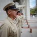 A group of U.S. Navy personnel in Service Khaki uniforms are standing in a formal line formation outdoors, likely during a ceremony or official event. The lead sailor in the foreground has his mouth open, appearing to be calling out a command or responding to one. The setting appears to be a public plaza or government building exterior.