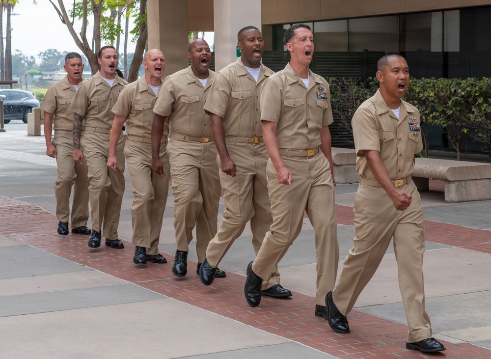 A group of approximately seven U.S. Navy personnel in Service Khaki uniforms are marching in formation on a brick-paved walkway, appearing to chant or call cadence in unison. They are moving in a coordinated column, suggesting a formal military drill or ceremonial march. The setting appears to be a military installation or government facility with palm trees and modern buildings visible in the background.