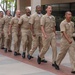 A group of approximately seven U.S. Navy personnel in Service Khaki uniforms are marching in formation on a brick-paved walkway, appearing to chant or call cadence in unison. They are moving in a coordinated column, suggesting a formal military drill or ceremonial march. The setting appears to be a military installation or government facility with palm trees and modern buildings visible in the background.