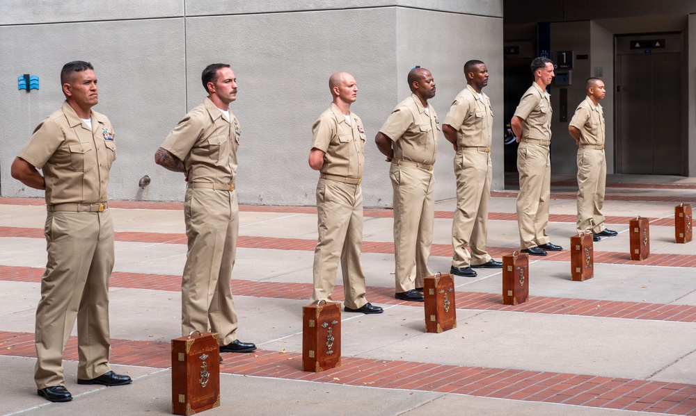 Seven U.S. Navy personnel in khaki service uniforms are standing at parade rest in a formal outdoor formation, each with a decorative wooden box placed at their feet. The setting appears to be a military installation courtyard or plaza with brick paving and a concrete building in the background. This appears to be a ceremonial event, likely a chief petty officer pinning or advancement ceremony.