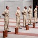 Seven U.S. Navy personnel in khaki service uniforms are standing at parade rest in a formal outdoor formation, each with a decorative wooden box placed at their feet. The setting appears to be a military installation courtyard or plaza with brick paving and a concrete building in the background. This appears to be a ceremonial event, likely a chief petty officer pinning or advancement ceremony.