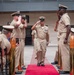 A formal U.S. Navy military ceremony is taking place outdoors, featuring a senior enlisted sailor walking down a red carpet aisle while fellow sailors render salutes on both sides. The setting appears to be a courtyard or outdoor plaza of a military building, with rows of seated attendees including civilians and uniformed personnel. The ceremony is consistent with a Chief Petty Officer pinning, retirement, or change of command event.