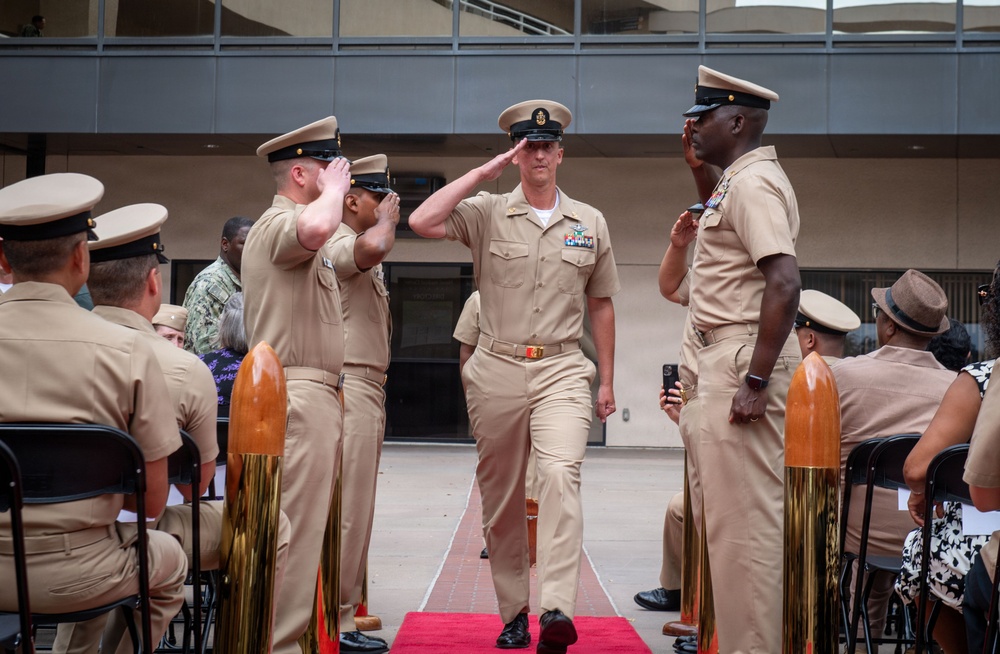 A U.S. Navy military ceremony is taking place outdoors in what appears to be a courtyard or plaza. A senior enlisted sailor in khaki service uniform walks down a red carpet while fellow sailors render salutes on both sides, suggesting a formal honors passage or retirement/promotion ceremony. Seated personnel and civilian guests observe the proceedings from chairs arranged on either side.
