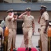 A U.S. Navy military ceremony is taking place outdoors in what appears to be a courtyard or plaza. A senior enlisted sailor in khaki service uniform walks down a red carpet while fellow sailors render salutes on both sides, suggesting a formal honors passage or retirement/promotion ceremony. Seated personnel and civilian guests observe the proceedings from chairs arranged on either side.