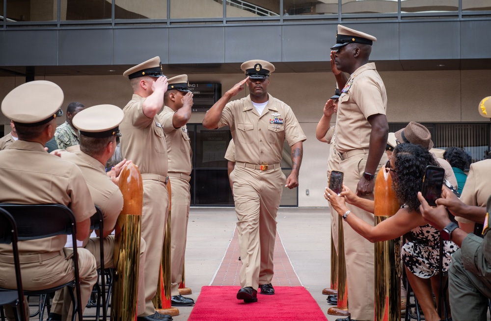 A U.S. Navy military ceremony is taking place outdoors, likely a promotion or retirement ceremony. A senior enlisted sailor in khaki service uniform walks down a red carpet flanked by gold stanchions while fellow sailors render salutes. Civilian guests and family members are seated and standing on the sides, recording the event on phones.