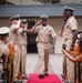 A U.S. Navy military ceremony is taking place outdoors, likely a promotion or retirement ceremony. A senior enlisted sailor in khaki service uniform walks down a red carpet flanked by gold stanchions while fellow sailors render salutes. Civilian guests and family members are seated and standing on the sides, recording the event on phones.