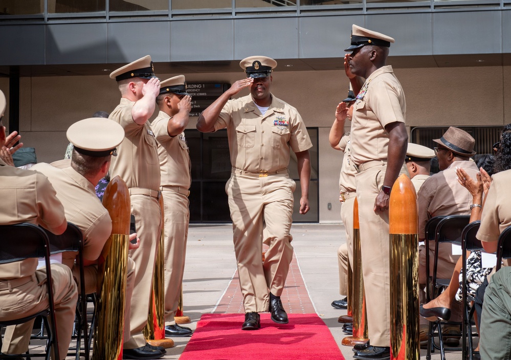 A formal U.S. Navy ceremony is taking place outdoors, likely a Chief Petty Officer pinning or promotion ceremony. Personnel in tan/khaki Service Uniform B are walking down a red carpet flanked by large brass shell casings used as ceremonial stanchions. Attendees seated on both sides are saluting the individuals proceeding down the aisle.