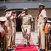 A formal U.S. Navy ceremony is taking place outdoors, likely a Chief Petty Officer pinning or promotion ceremony. Personnel in tan/khaki Service Uniform B are walking down a red carpet flanked by large brass shell casings used as ceremonial stanchions. Attendees seated on both sides are saluting the individuals proceeding down the aisle.