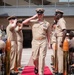 A formal U.S. Navy military ceremony is taking place outdoors, featuring personnel in Service Khaki uniforms walking a red carpet flanked by decorative artillery shell stanchions. The central figure, a senior enlisted sailor wearing ribbons and a warfare device, is being rendered salutes by fellow sailors on both sides. Seated civilians and uniformed personnel observe the ceremony from folding chairs on either side.