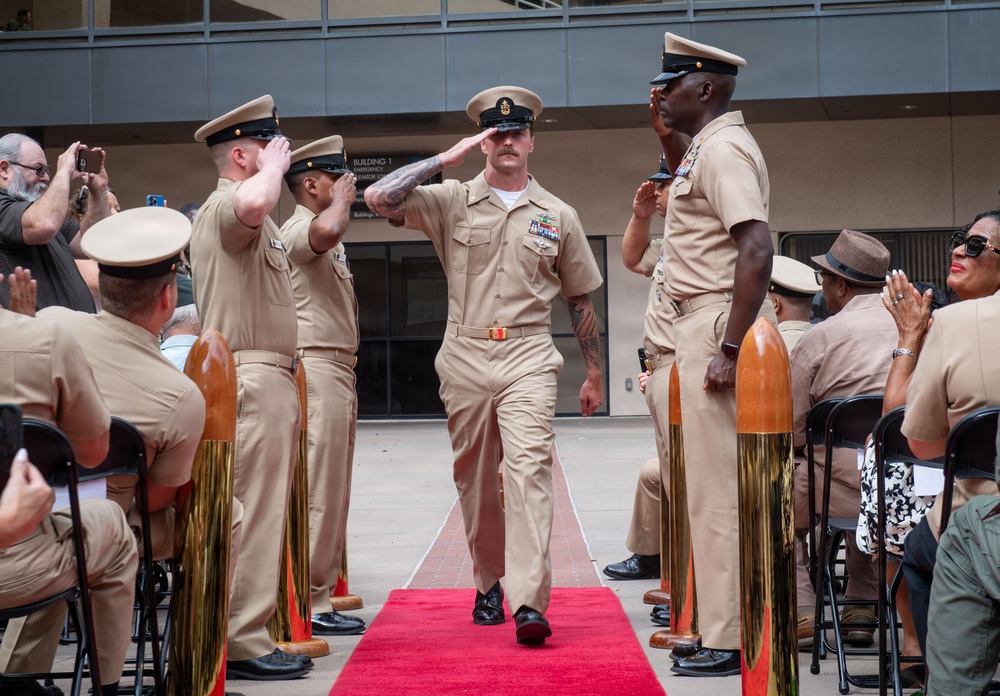 A U.S. Navy Chief Petty Officer promotion or retirement ceremony is taking place outdoors at what appears to be a naval installation. The central figure walks down a red carpet flanked by saluting personnel and decorative shell casing bollards, while seated guests and standing sailors observe and applaud. The formal ceremony follows traditional Navy Chief's Mess traditions.
