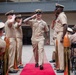 A U.S. Navy Chief Petty Officer promotion or retirement ceremony is taking place outdoors at what appears to be a naval installation. The central figure walks down a red carpet flanked by saluting personnel and decorative shell casing bollards, while seated guests and standing sailors observe and applaud. The formal ceremony follows traditional Navy Chief's Mess traditions.