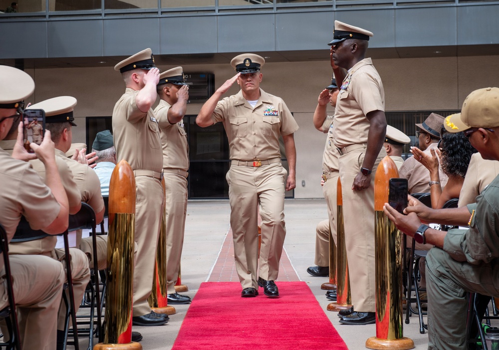 A U.S. Navy military ceremony is taking place outdoors, featuring a senior enlisted sailor walking down a red carpet aisle while fellow sailors render salutes on both sides. The scene appears to be a formal retirement or change of command ceremony, indicated by the ceremonial decorations including gold shell casings on wooden pedestals flanking the red carpet. Seated attendees, including civilians and uniformed personnel, observe and photograph the event.
