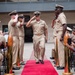 A U.S. Navy military ceremony is taking place outdoors, featuring a senior enlisted sailor walking down a red carpet aisle while fellow sailors render salutes on both sides. The scene appears to be a formal retirement or change of command ceremony, indicated by the ceremonial decorations including gold shell casings on wooden pedestals flanking the red carpet. Seated attendees, including civilians and uniformed personnel, observe and photograph the event.