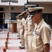 A formal military ceremony is taking place outside a Naval Medical Center, with a line of U.S. Navy personnel standing at attention in Service Khaki uniforms. The personnel are aligned in a row with wooden presentation cases placed on the ground beside them, suggesting an awards or promotion ceremony. A podium bearing an official seal is visible in the background.