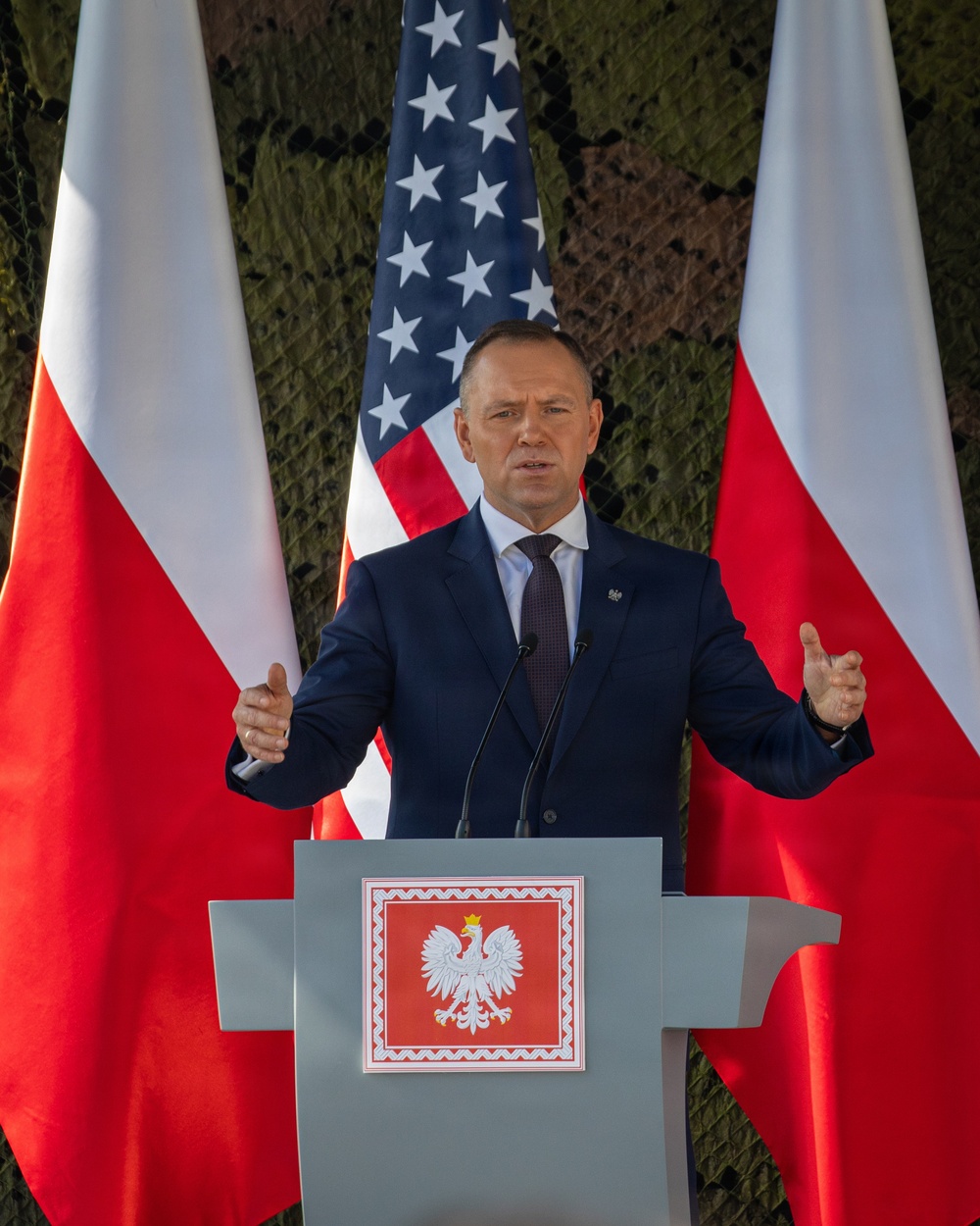 A man in a dark navy suit is speaking at a podium bearing the coat of arms of Poland (white eagle on red background). The setting features two Polish flags flanking the speaker and a United States flag centered behind him, against a military camouflage netting backdrop. The formal arrangement suggests an official bilateral or diplomatic/military event.